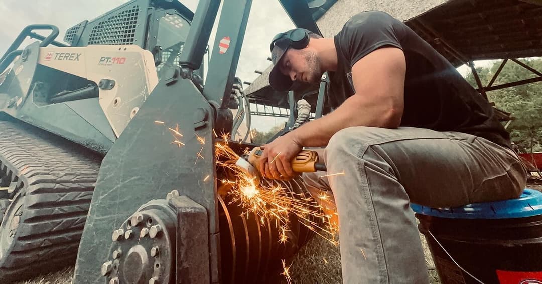 Man welding on a Terex skid steer loader, sparks flying, in an outdoor workshop setting.