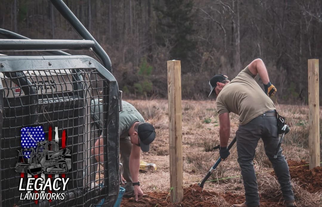 Two workers digging and installing posts for a fence in a clearing, with equipment nearby.