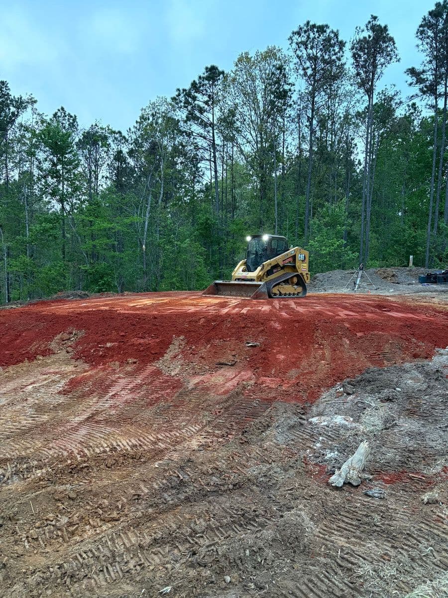 Bulldozer leveling red dirt on a construction site surrounded by trees.