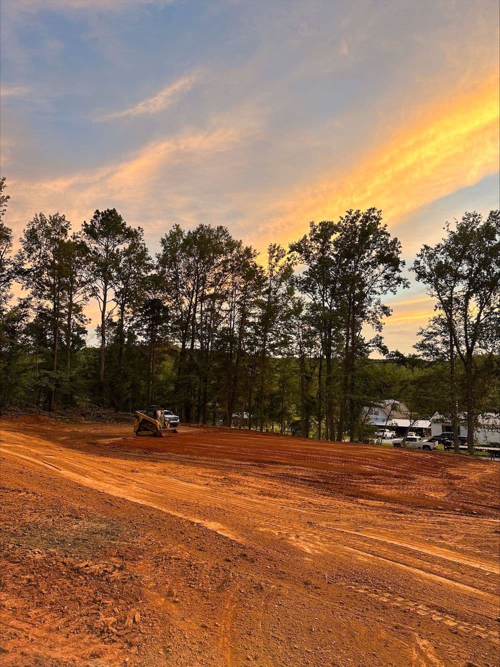 Cleared land with construction equipment at sunset, surrounded by trees and a colorful sky.