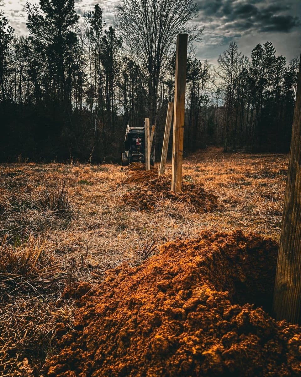 Construction site with heavy machinery and fence posts in a wooded area during sunset.