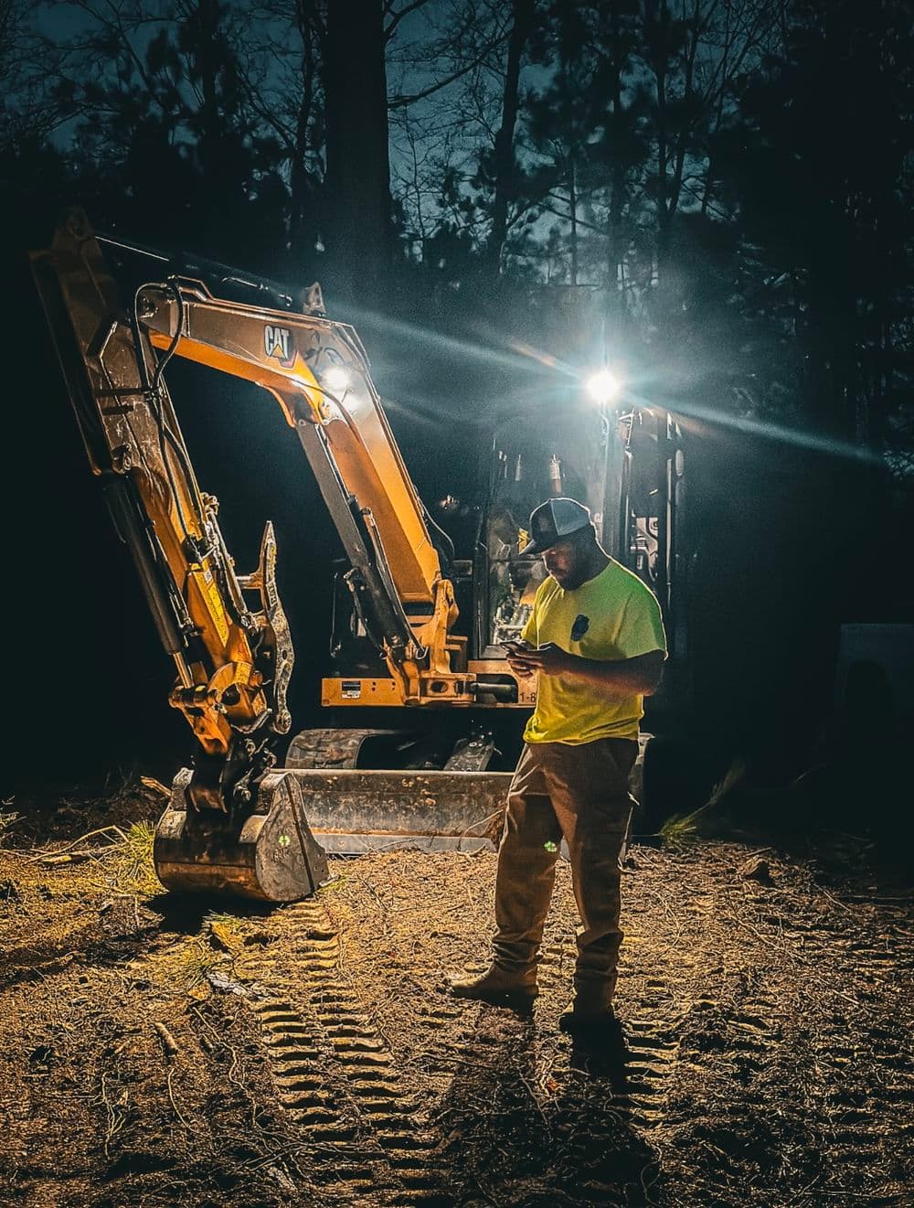 Construction worker in neon shirt operating CAT excavator at night in forest clearing.