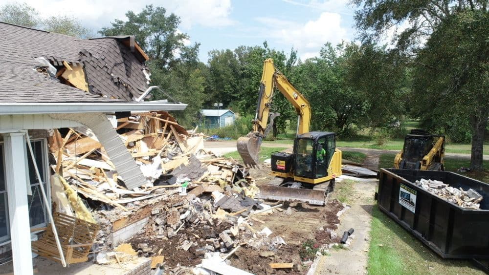 Construction equipment demolishing a damaged home, debris scattered around, clear skies.