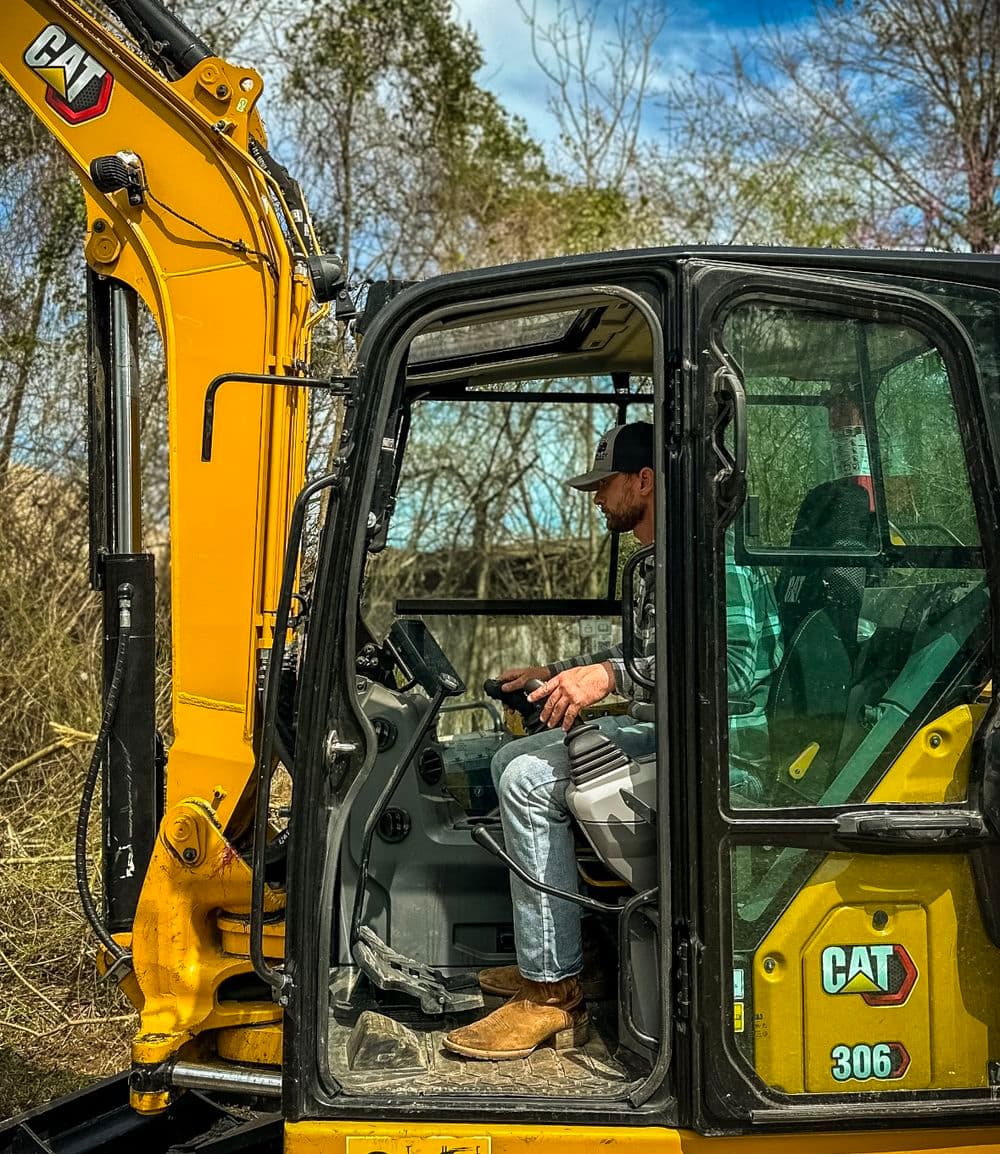 Operator inside a CAT 306 excavator, focused on controls amidst a natural backdrop.