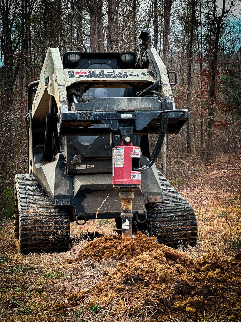 Heavy-duty skid steer loader digging in wooded area with freshly disturbed soil.