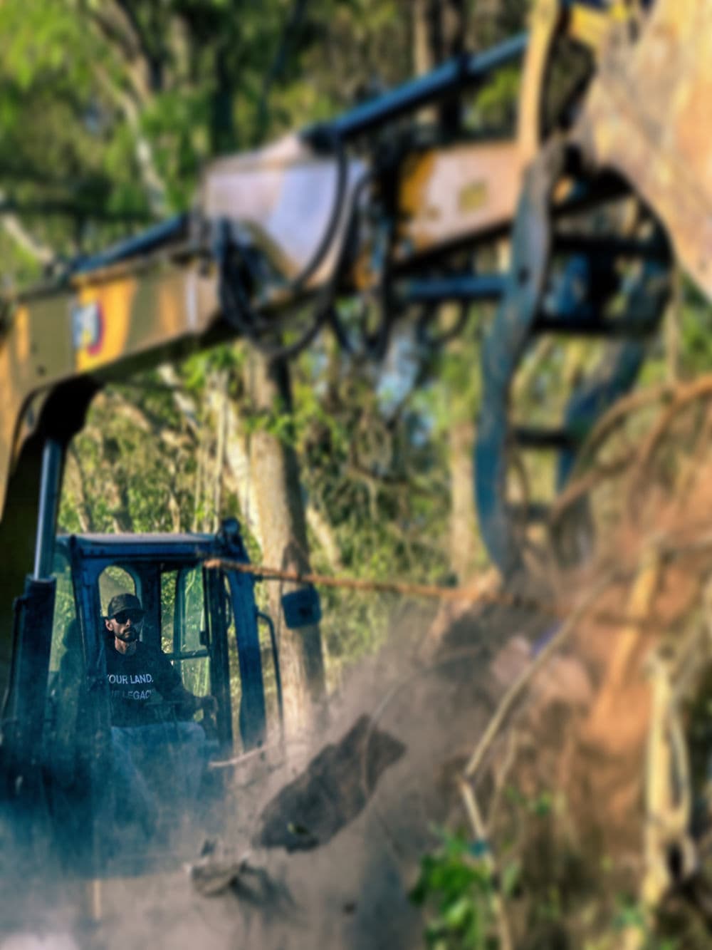 Operator using heavy machinery to clear trees in a forested area. Dust and debris visible.