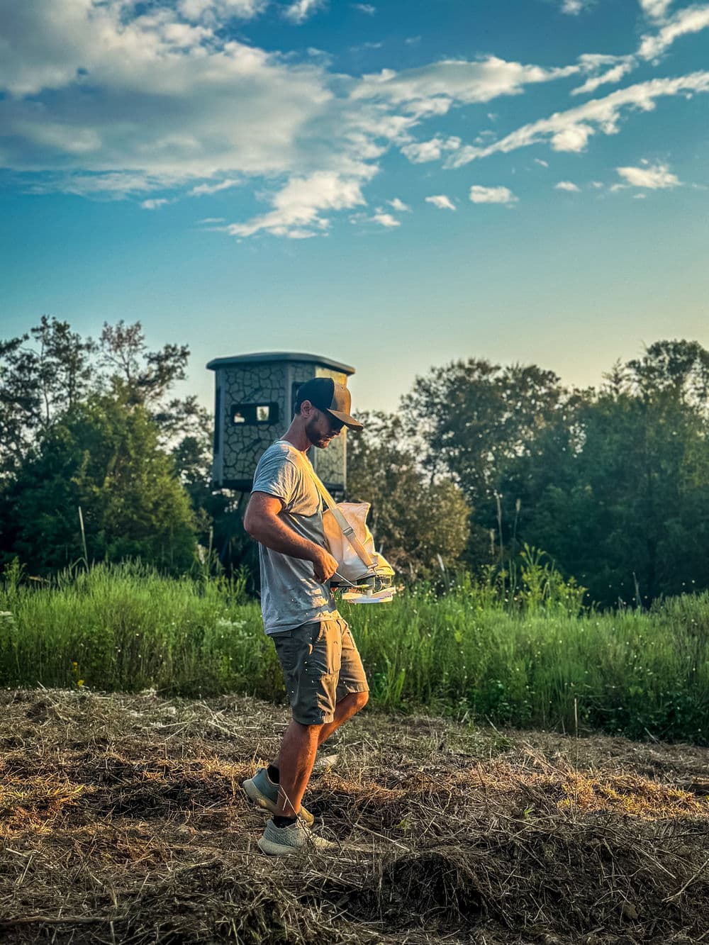 Man carrying a hunting backpack near a wildlife observation tower at sunset.