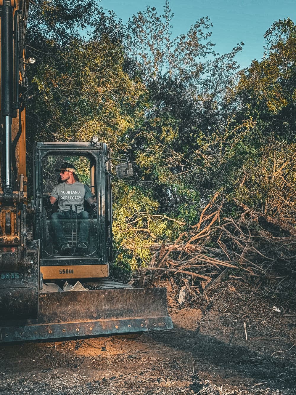 Excavator operator clearing land, surrounded by greenery and piled brush in a construction site.