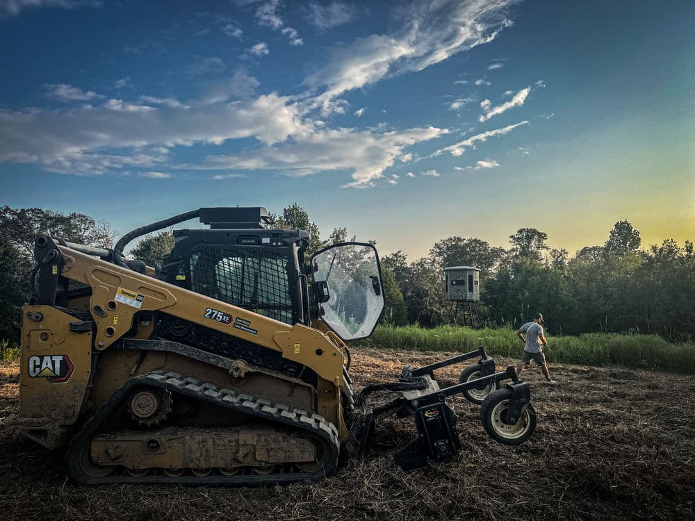 Skid steer loader with tracks in a field at sunset, with a person in the background.