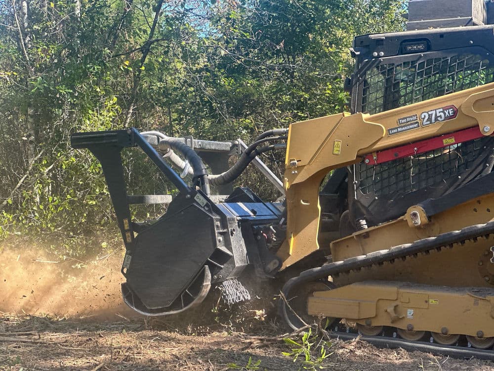 Stump grinder attachment on heavy machinery clearing woodland vegetation and dirt.
