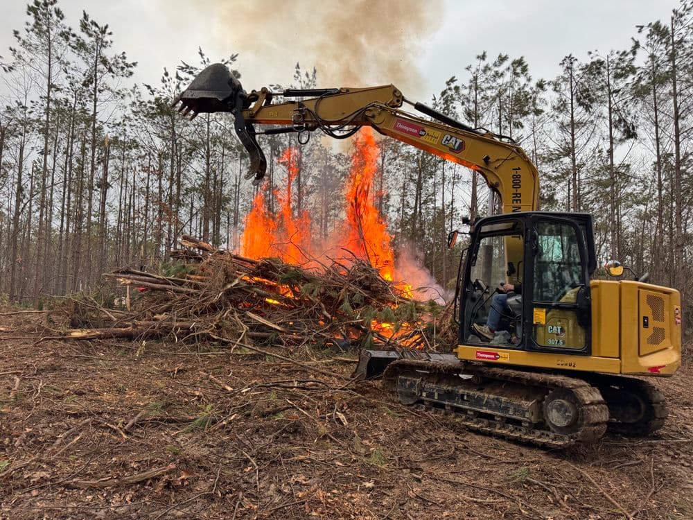 Excavator operator burning tree debris in a forest clearing with flames and smoke.