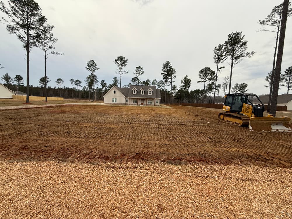 Newly cleared residential lot with a house in the background and construction equipment in view.