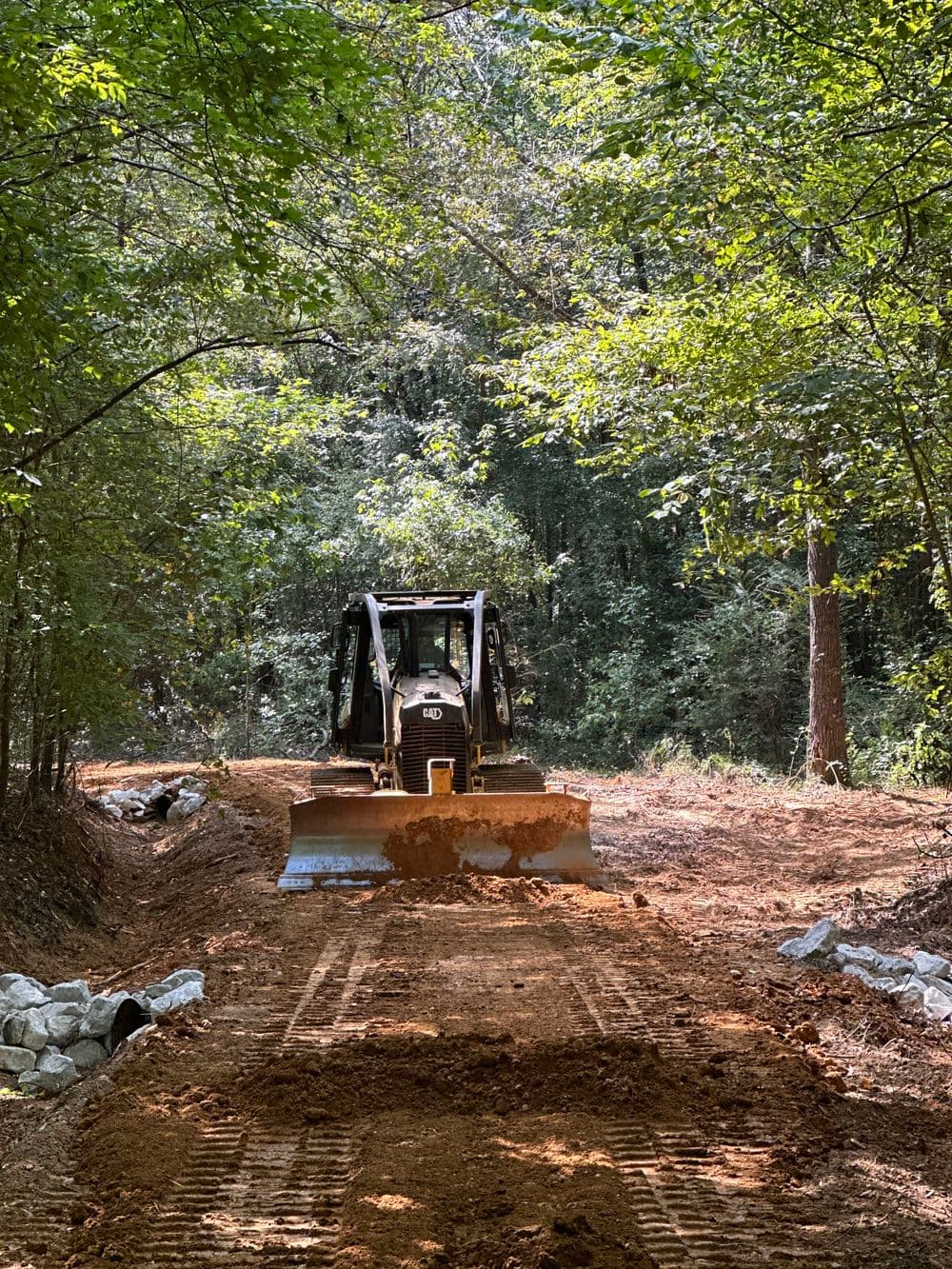 Bulldozer clearing a dirt road through a wooded area, surrounded by green trees.